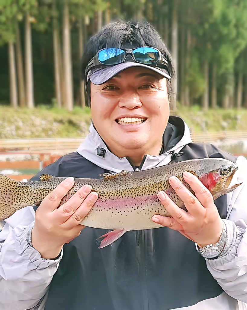 むっちぃ（西川睦雄）顔写真。
趣ののエリアトラウトでビッグサイズのニジマスを釣り上げてスマイルの図。